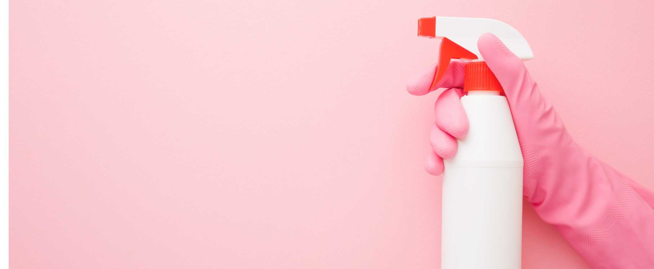Hand in rubber protective glove holding white spray bottle. Detergent for different surfaces in kitchen, bathroom and other rooms. Closeup. Empty place for text or logo. Light pastel pink background.
