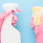 Hands in rubber protective gloves holding white spray bottle and foamy sponge. Detergent for different surfaces in kitchen, bathroom and other rooms. Pastel blue background. Point of view shot.