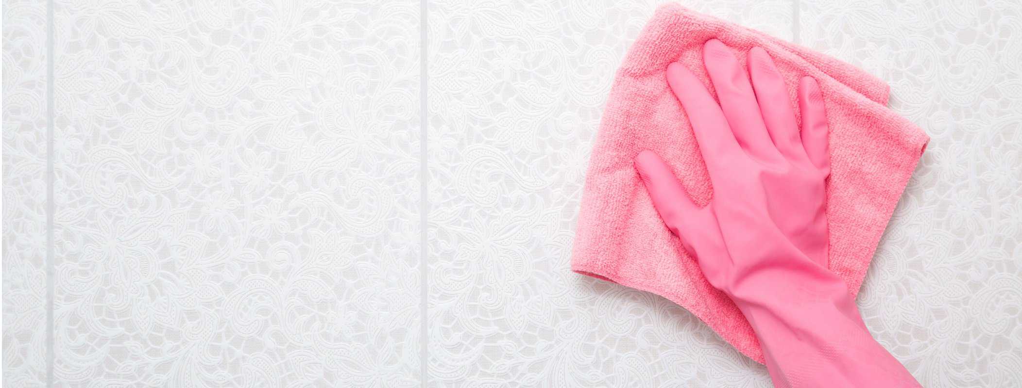 Woman hand in pink rubber protective glove wiping light gray ceramic tiles wall with dry rag. Closeup. Spring general or regular cleanup. Empty place for text or logo.
