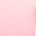 Young adult woman hand holding white spray bottle. Detergent for different surfaces in kitchen, bathroom and other rooms. Closeup. Empty place for text or logo. Light pink background. Pastel color.