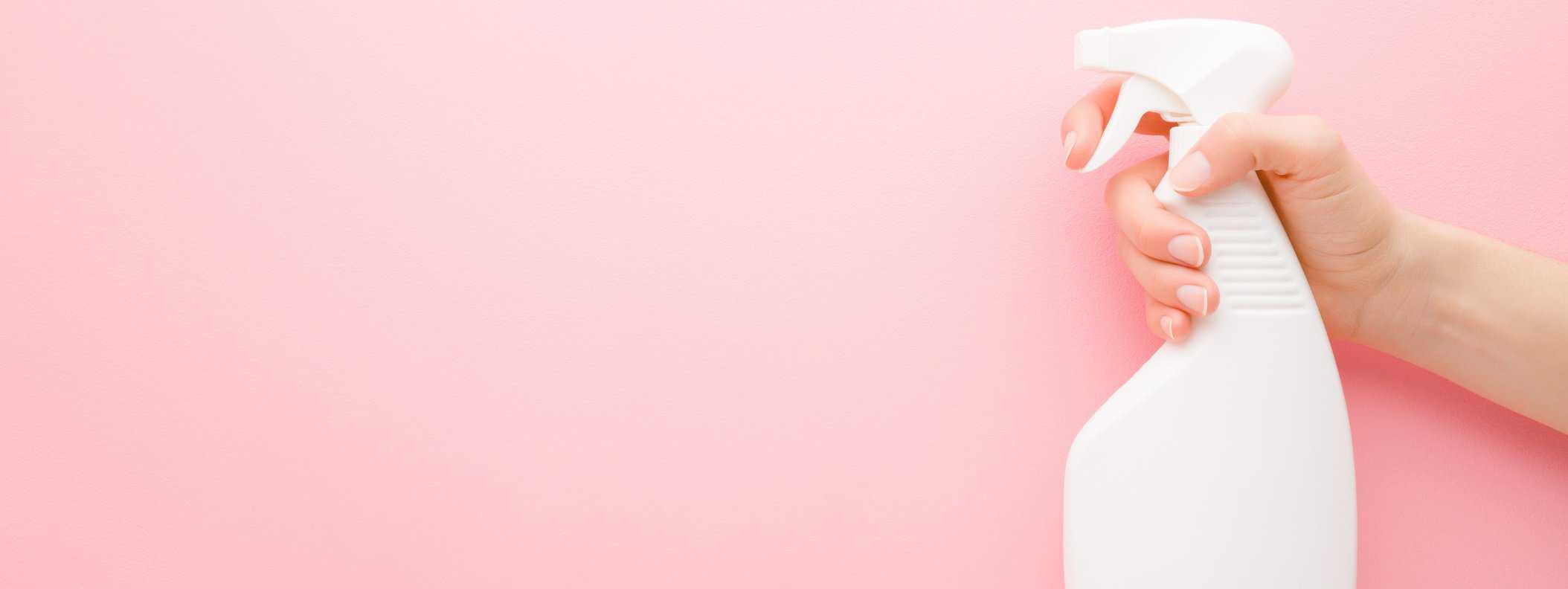 Young adult woman hand holding white spray bottle. Detergent for different surfaces in kitchen, bathroom and other rooms. Closeup. Empty place for text or logo. Light pink background. Pastel color.
